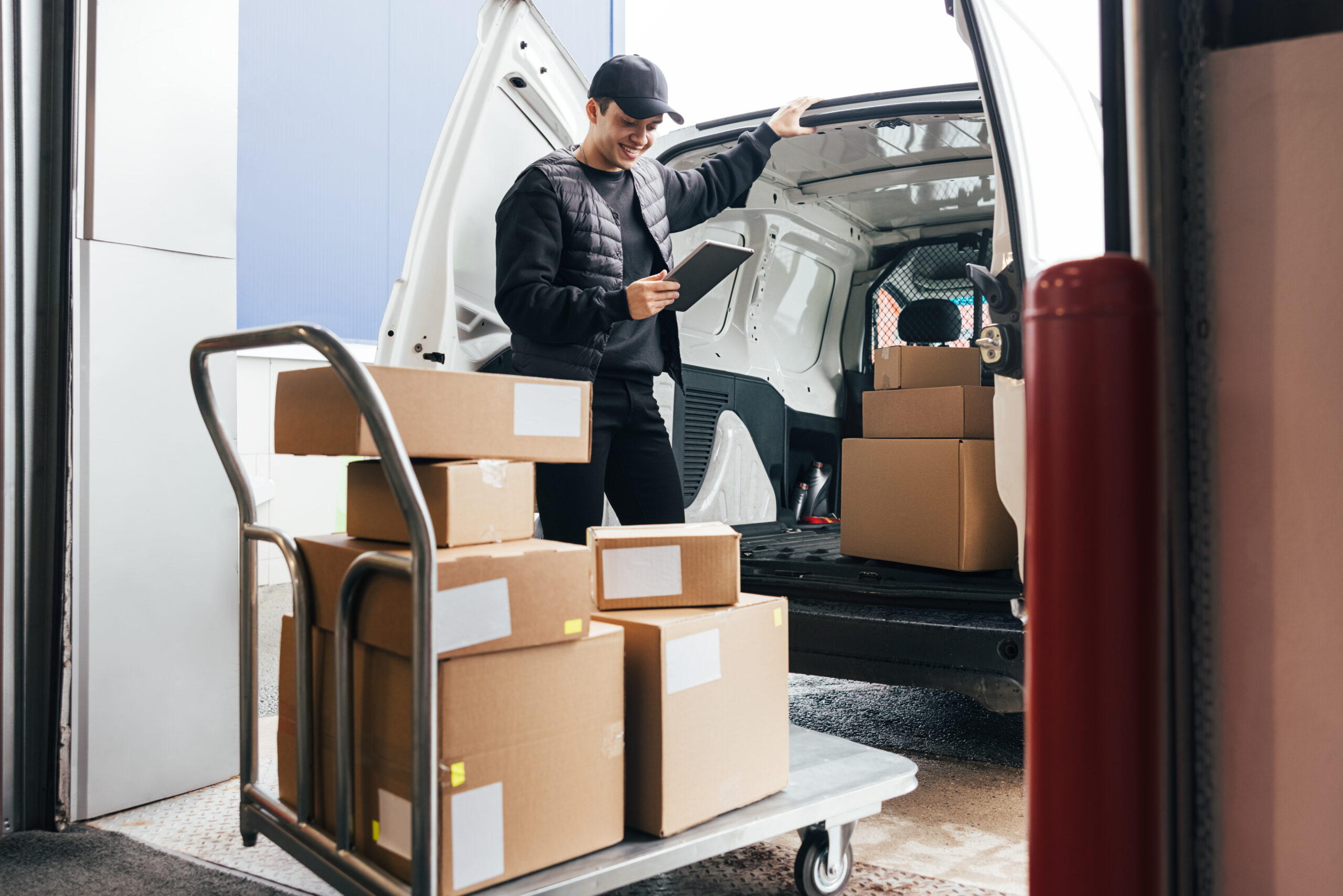 Courier in uniform holding a digital tablet checking amount of packages on a cart while standing at a car